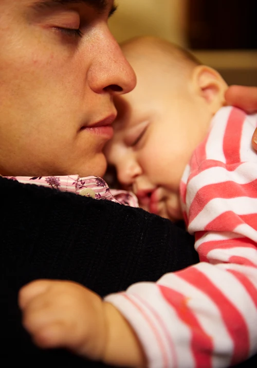 Dad sleeps seated as he holds his sleeping baby.