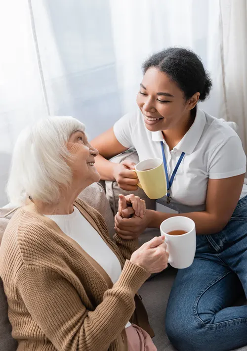 A young female caregiver having coffee at home with an older woman.