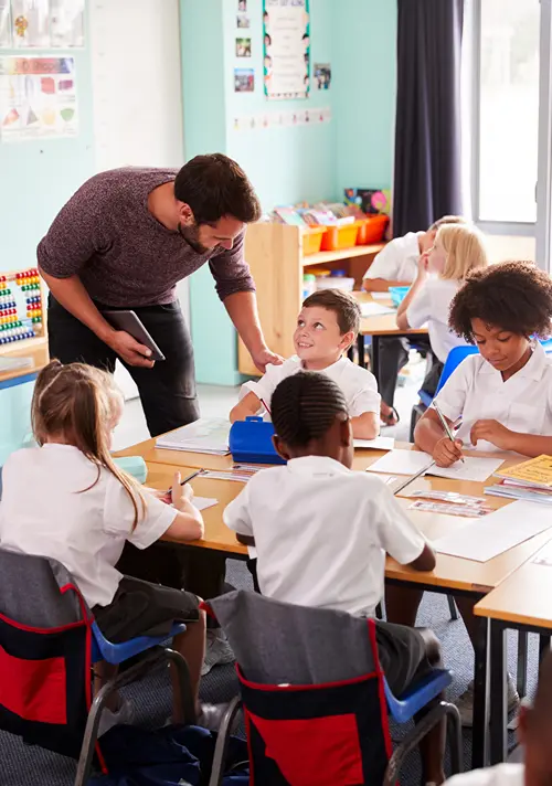 A teacher bends down to speak to a group of students seated at a classroom work table.