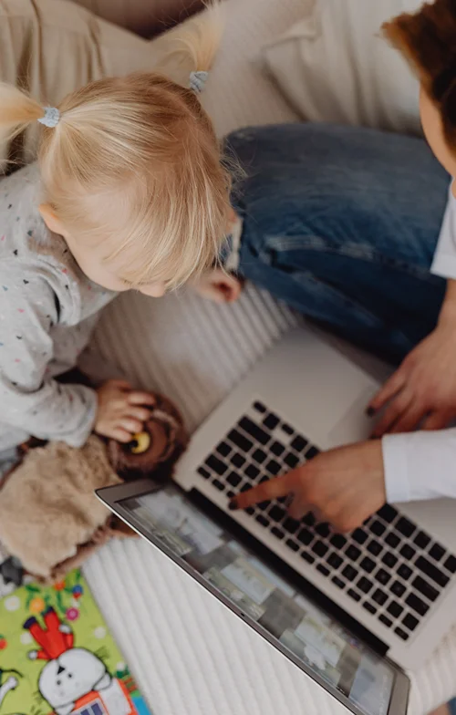 Overhead shot of a parent using a mac laptop while a young child with a stuffed animal watches.