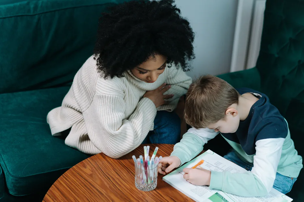 An in-home tutor helping an elementary student complete a homework assignment at a table with a cup of pens.