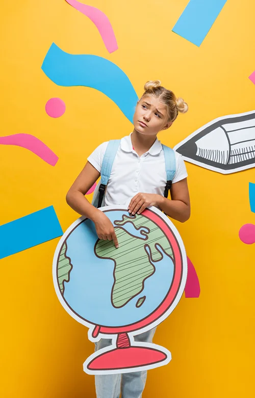 A female teenage student stands against a yellow background, holding and pointing at a large, flat paper cutout of a globe.