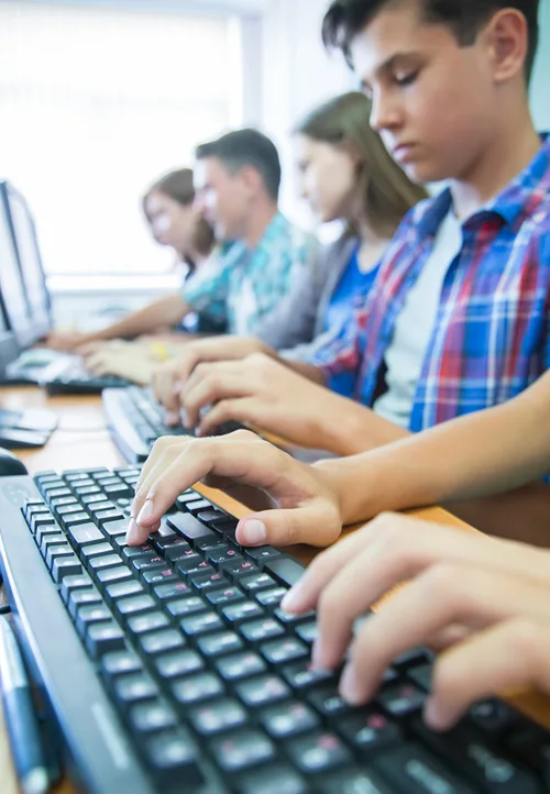 A row of students typing on black computer keyboards in a classroom, with the focus on a student's hands in the foreground.
