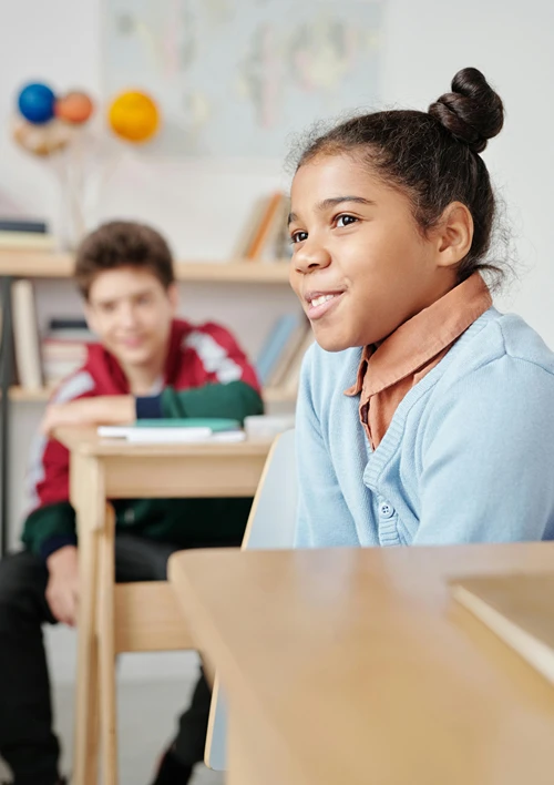 A middle school classroom showing a girl smiling off to the side while a boy watched from his desk behind her.