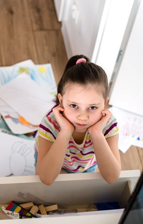 A young girl in a messy room looks up with a frown as she rests her elbows on an open dresser drawer.