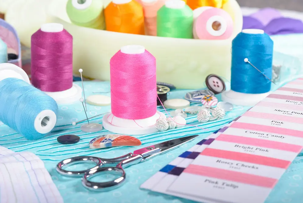 Pink, purple, and blue sewing thread spools, scissors, and a pink color swatch on a light blue fabric background.