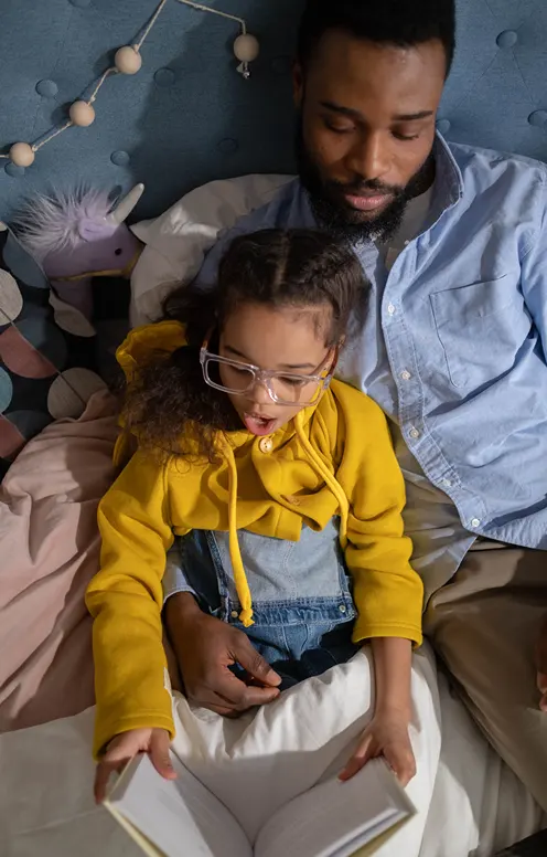 A little girl with glasses reading a book on dad's lap.