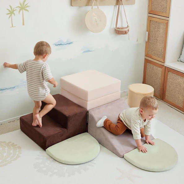 Two toddlers playing on a set of soft, neutral-colored foam climbing blocks and a ramp in a playroom.