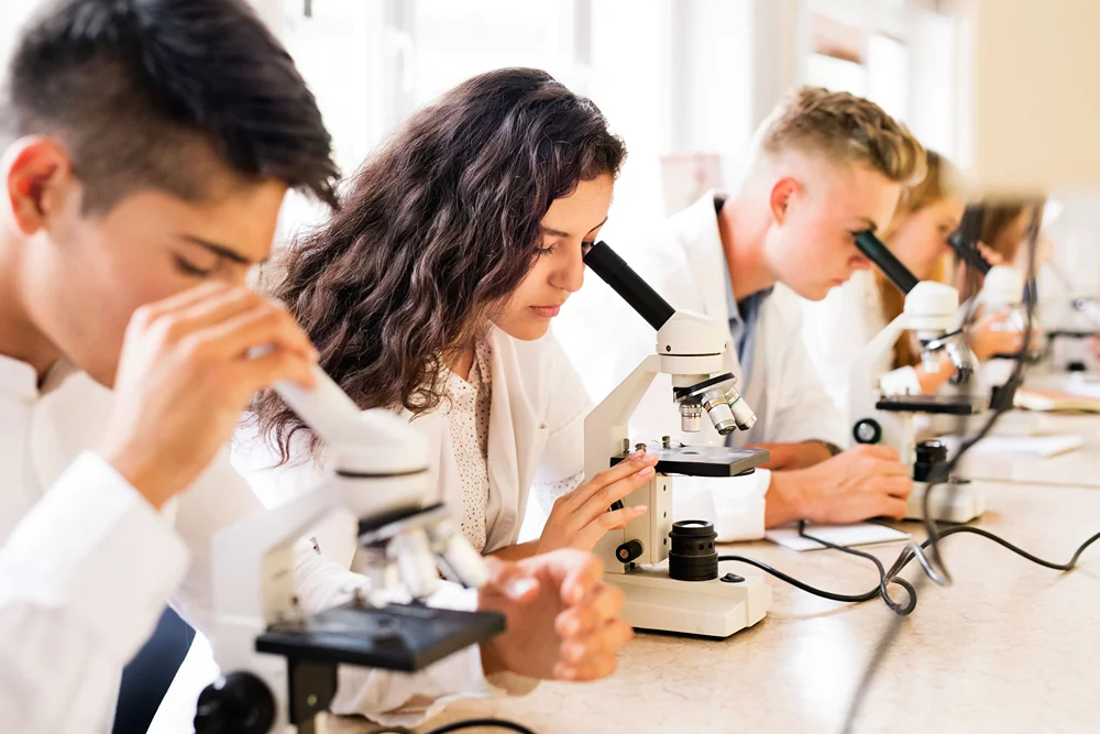 A group of teenage students in white lab coats sitting at a long table and looking into microscopes during a science class..