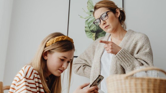 A mother points her finger at her daughter who looks distressed while looking at her phone.
