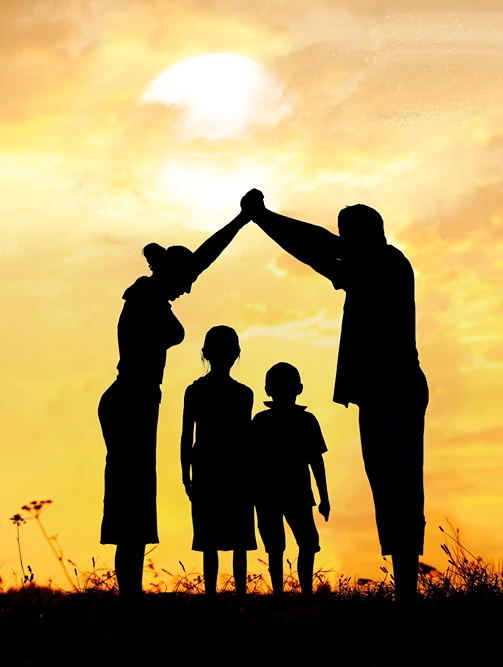 Silhouette of a mother and father forming a protective roof shape with their arms over their two children during a golden sunset.