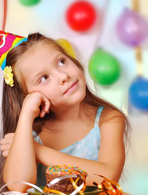 A young girl with party hat on her head and cake in her plate smiles and looks up at the balloons around her.