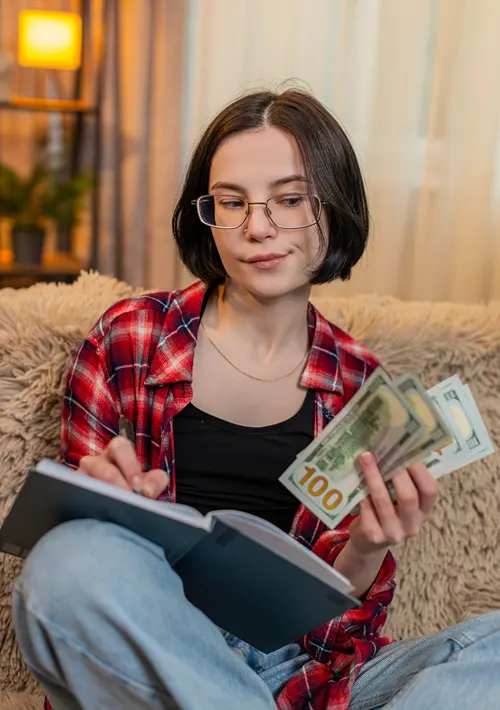 A teen girl holding cash money in one hand as she writes in a book ledger.