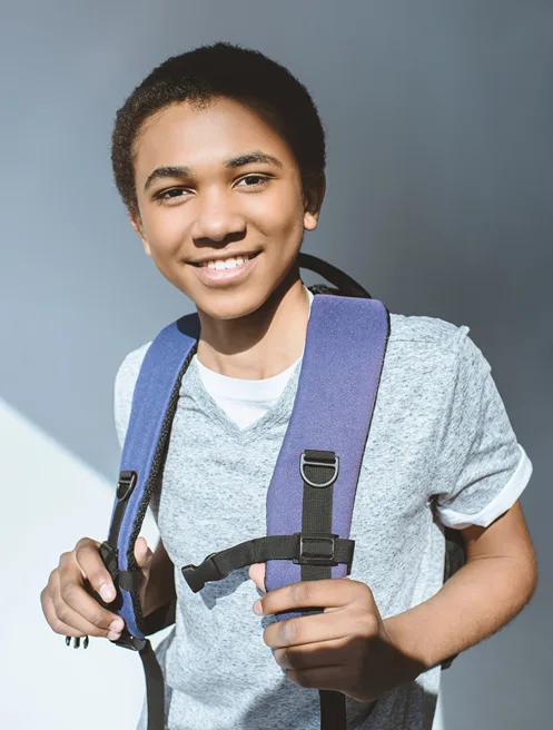Smiling teenage boy in a grey t-shirt holding the straps of his purple school backpack.