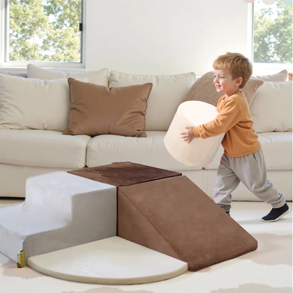 A young boy carrying a cylindrical foam block towards a set of soft climbing stairs and a ramp in a living room.