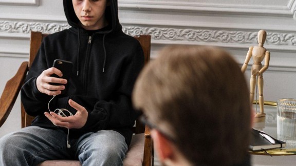 A tween in a black hoodie fiddles with his phone while listening to a therapist.