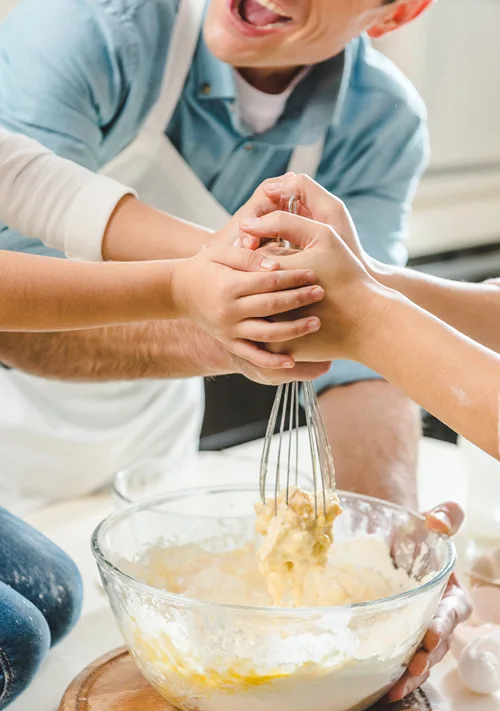Dad vigorously stirring cookie dough with a whisk as family members place there hands on his to help.
