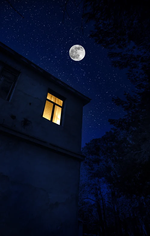 A lone lit room at night in the second story of a home under a full moon.