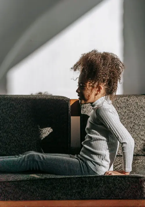 A young Black girl with curly hair sits cross-legged on a textured floor pad in a children's therapy clinic, surrounded by soft seating blocks, looking off to the side in natural light.