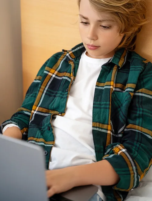 A young teen boy casually sits back while working on his laptop.