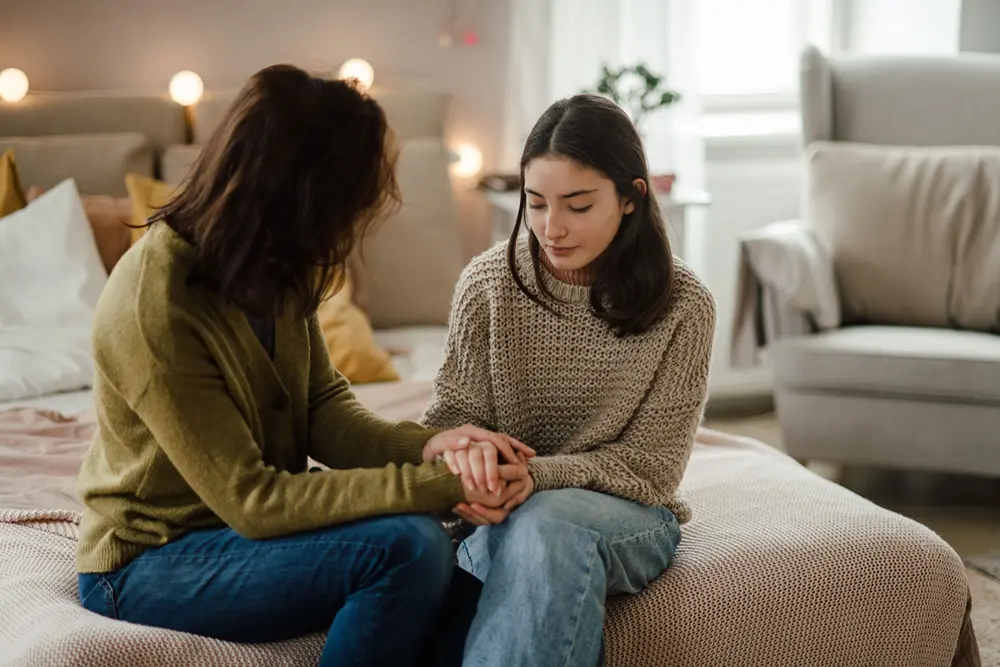 A parent and their child sitting at the foot of a bed holding hands. They appear to be having a serious conversation.