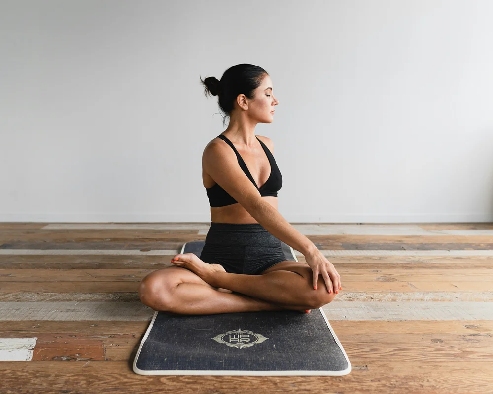 A woman sits cross legged on a yoga matt stretches her upper body and head to her left.