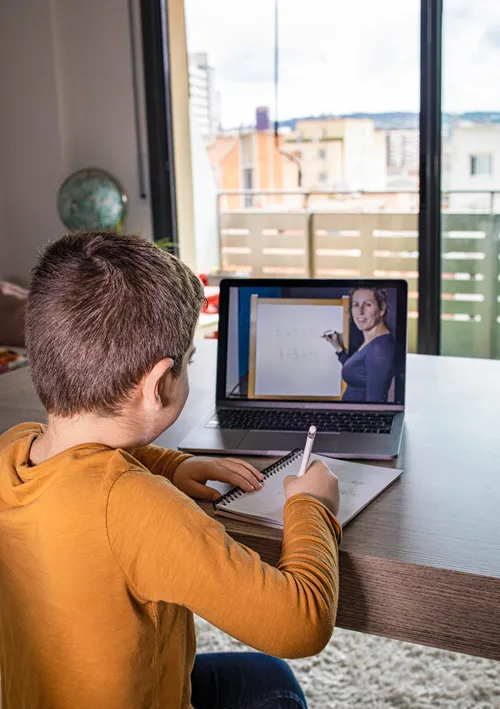 A teacher is shown on a laptop as a young boy is taking notes at home.
