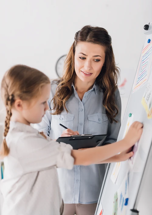 A tween girl works on a whiteboard as her therapist holds a clip board.