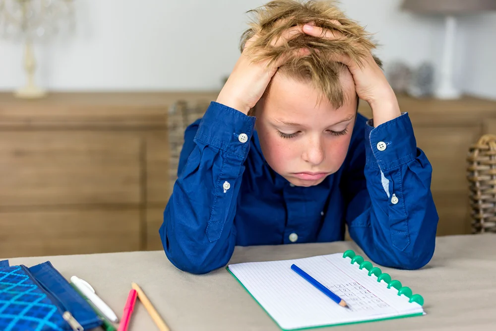 A young boy grabs his hair in frustration as he looks down at math equations on a notepad.