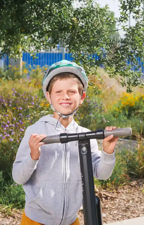 A boy wearing a helmet smiles as he stands beside his electric scooter.