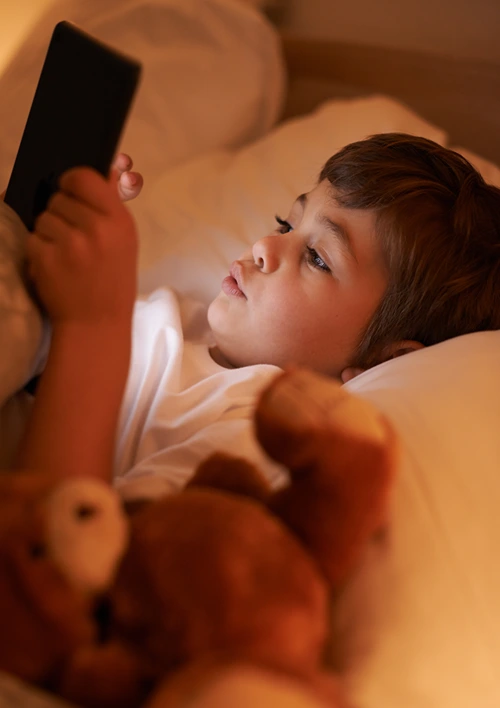 Boy looking at tablet in bed with teddy bear in dimly bedroom.