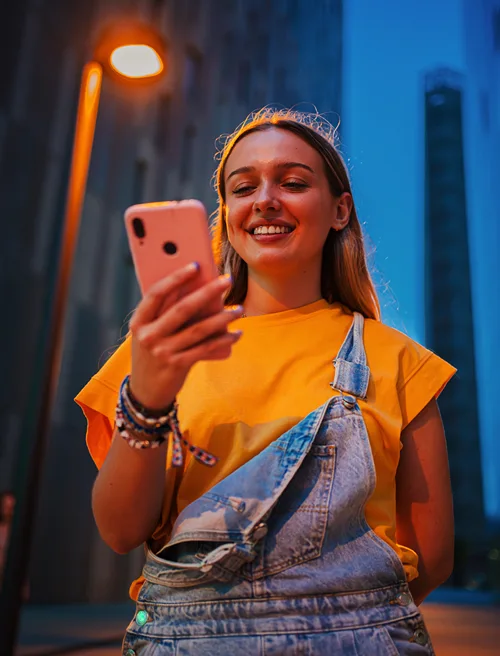 A young woman stands under a street lamp at night smiling at her iPhone.
