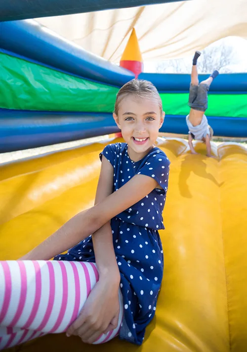 A smiling girl plays in a bouncy castle while a boy does a handstand in the background.