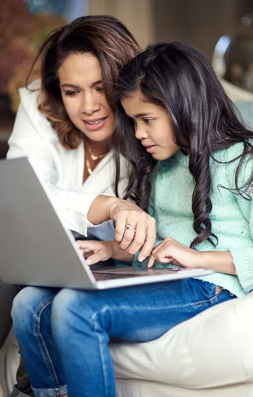 A mother helps her daughter use a laptop while seated on a sofa.