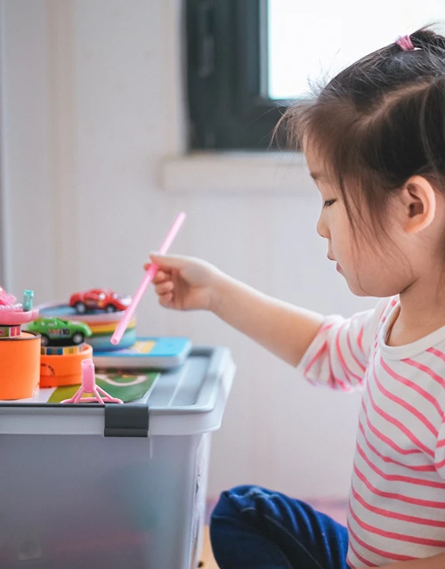 An Asian child in a striped shirt playing with toy cars and a pink straw next to a storage bin.