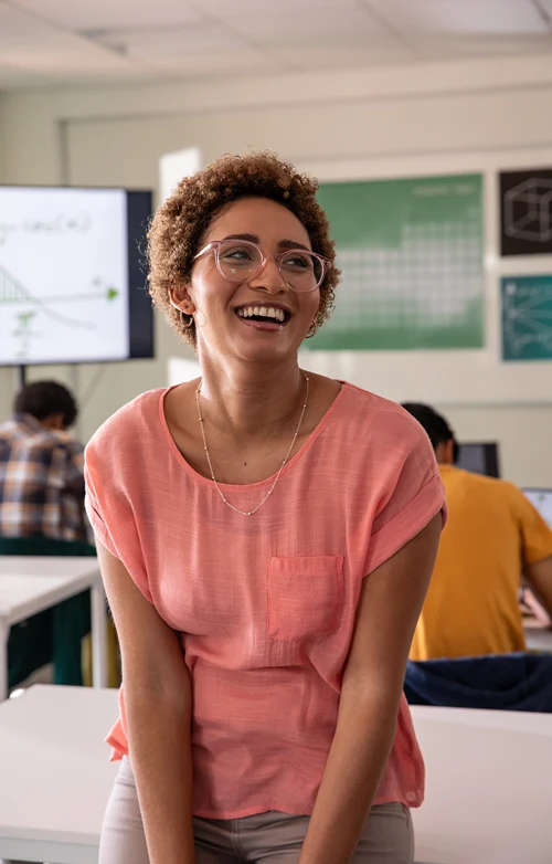 Female teacher leans against a school desk and smiles has her students work in the background.