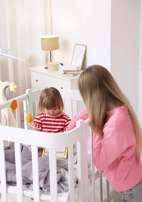 A female tween babysitter cares for a young child standing up in a crib.