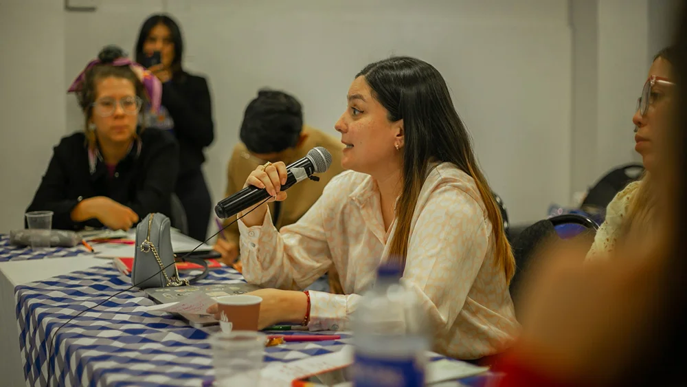 A woman with long dark hair and a cream patterned blouse speaks into a handheld microphone at a table while other attendees listen in the background.