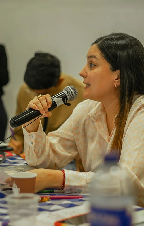 A woman sits at a table holding a mic and addresses attendees at public hall meeting.